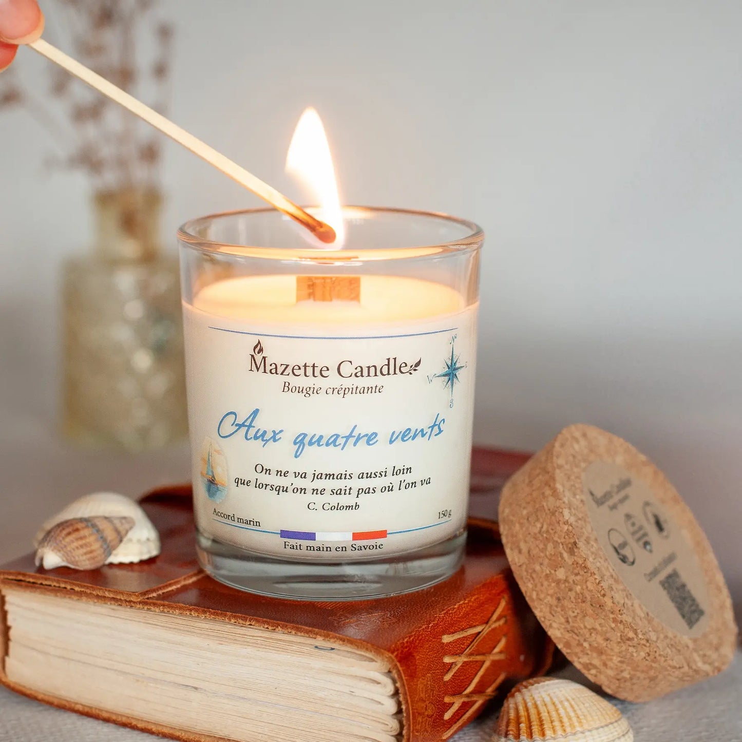 Bougie Mazette Candle Aux quatre vents 150g parfum accord marin, en cours d’allumage avec une allumette, couvercle en liège