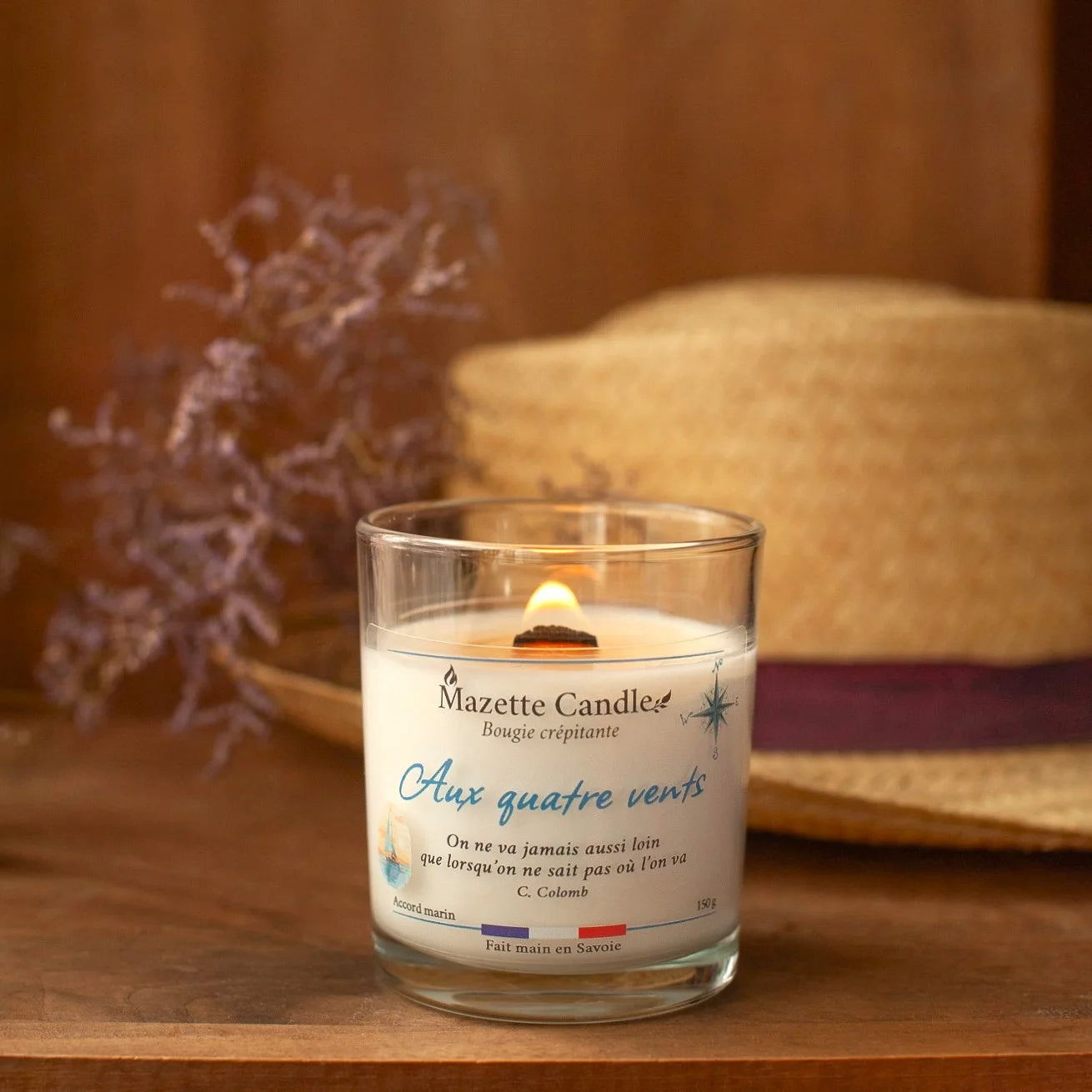 Bougie Mazette Candle Aux quatre vents 150g parfum accord marin, allumée devant un chapeau de paille et des fleurs séchées