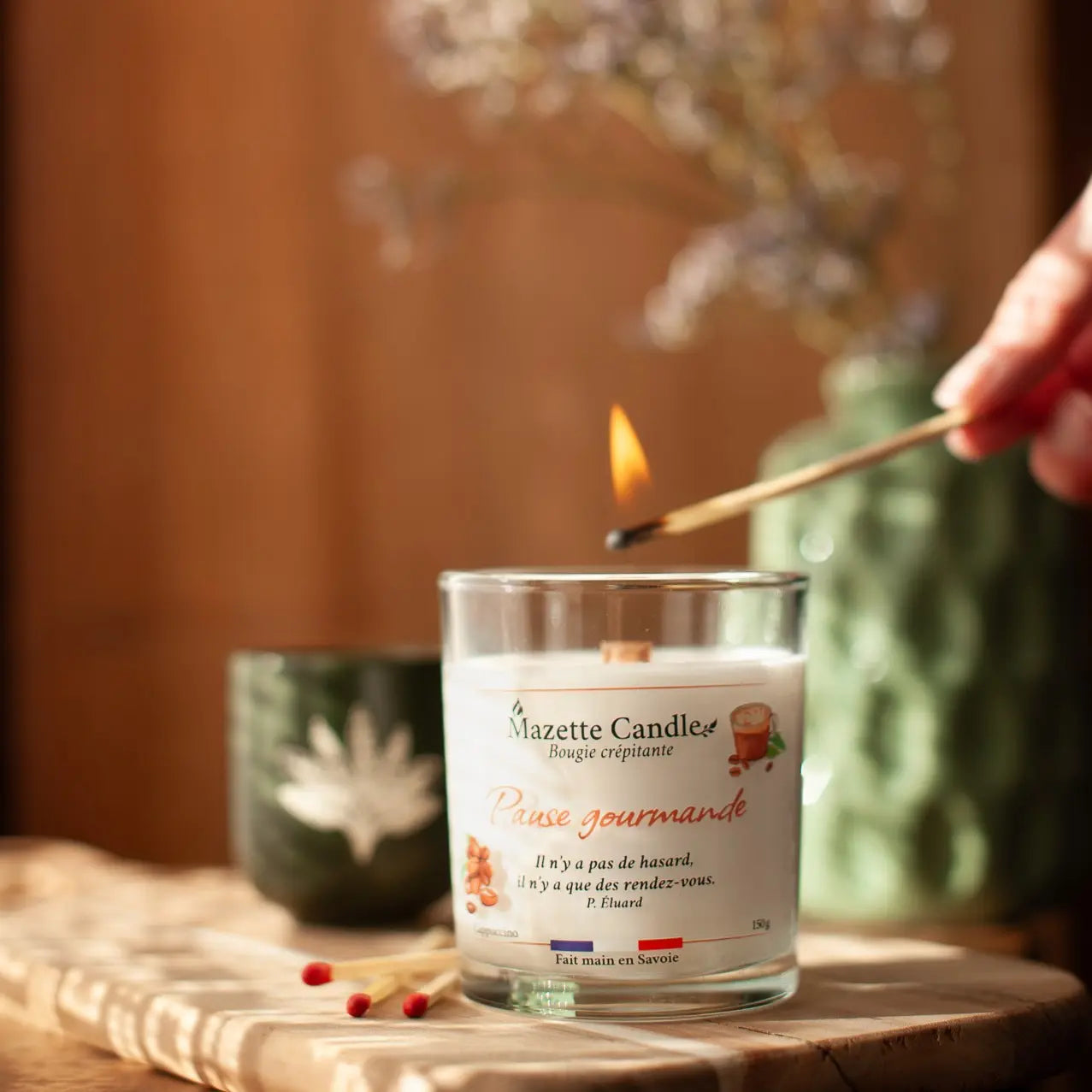 Bougie Mazette Candle Pause gourmande 150g parfum cappuccino, en cours d’allumage avec une allumette, tasse et vase en décor