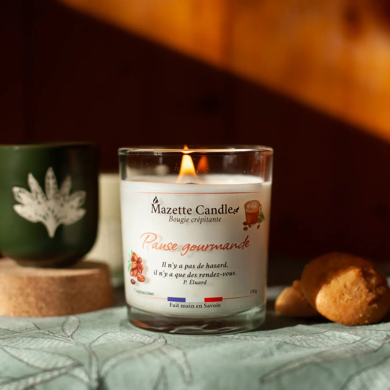 Bougie Mazette Candle Pause gourmande 150g parfum cappuccino, allumée sur une table, avec une tasse et des biscuits en décor
