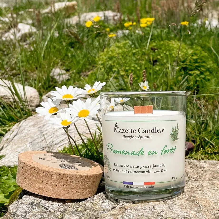 Bougie Mazette Candle Promenade en forêt 150g parfum cèdre, sur un rocher entouré de fleurs, son couvercle en liège à côté