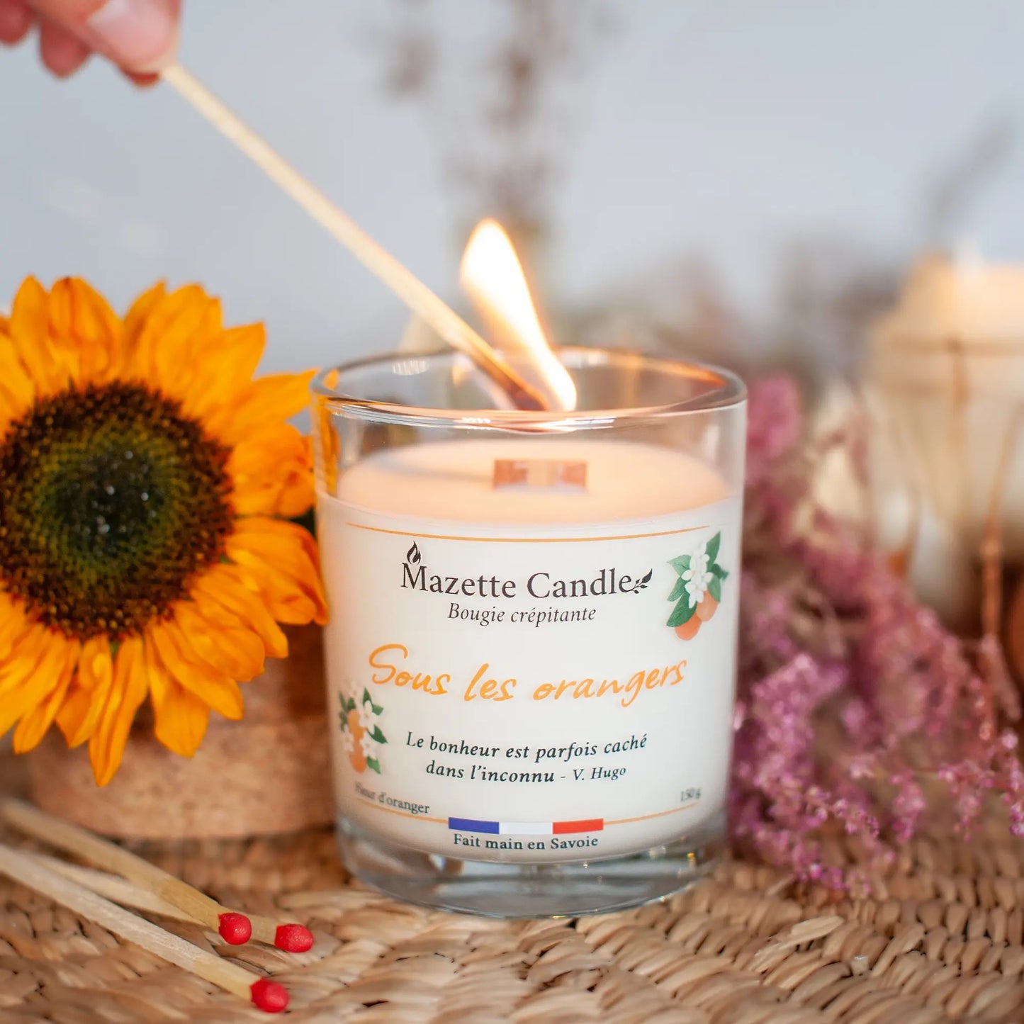 Bougie Mazette Candle Sous les orangers 150g parfum fleur d’oranger, en cours d’allumage avec une allumette, décor floral