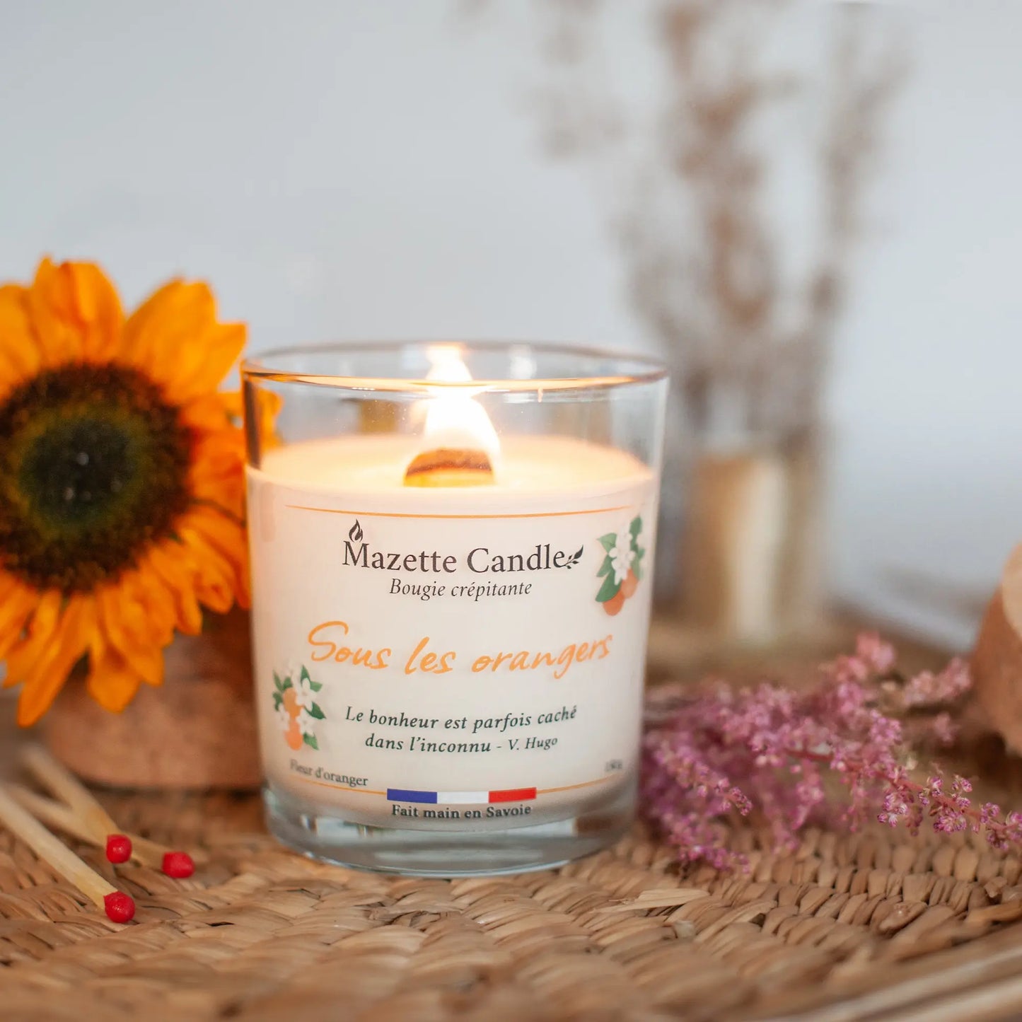 Bougie Mazette Candle Sous les orangers 150g parfum fleur d’oranger, allumée sur un plateau en osier dans un décor floral