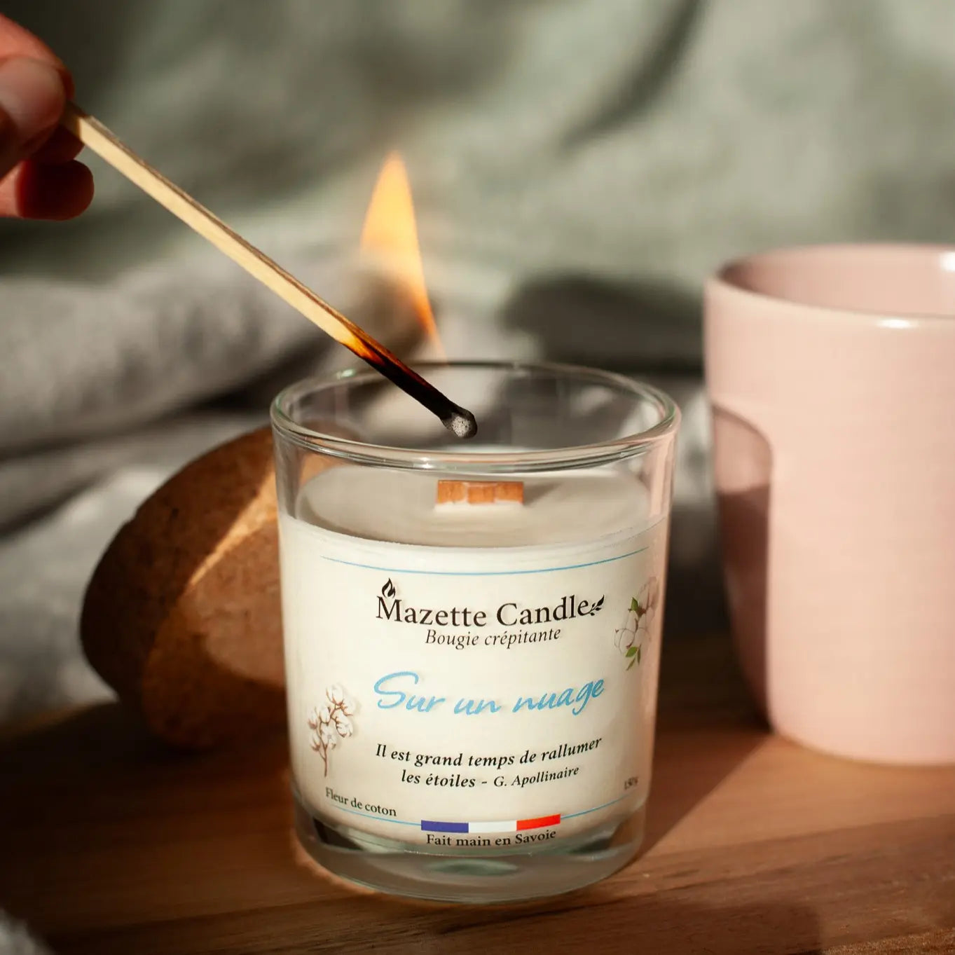 Bougie Mazette Candle Sur un nuage 150g parfum fleur de coton, en cours d’allumage avec une allumette, couvercle en liège