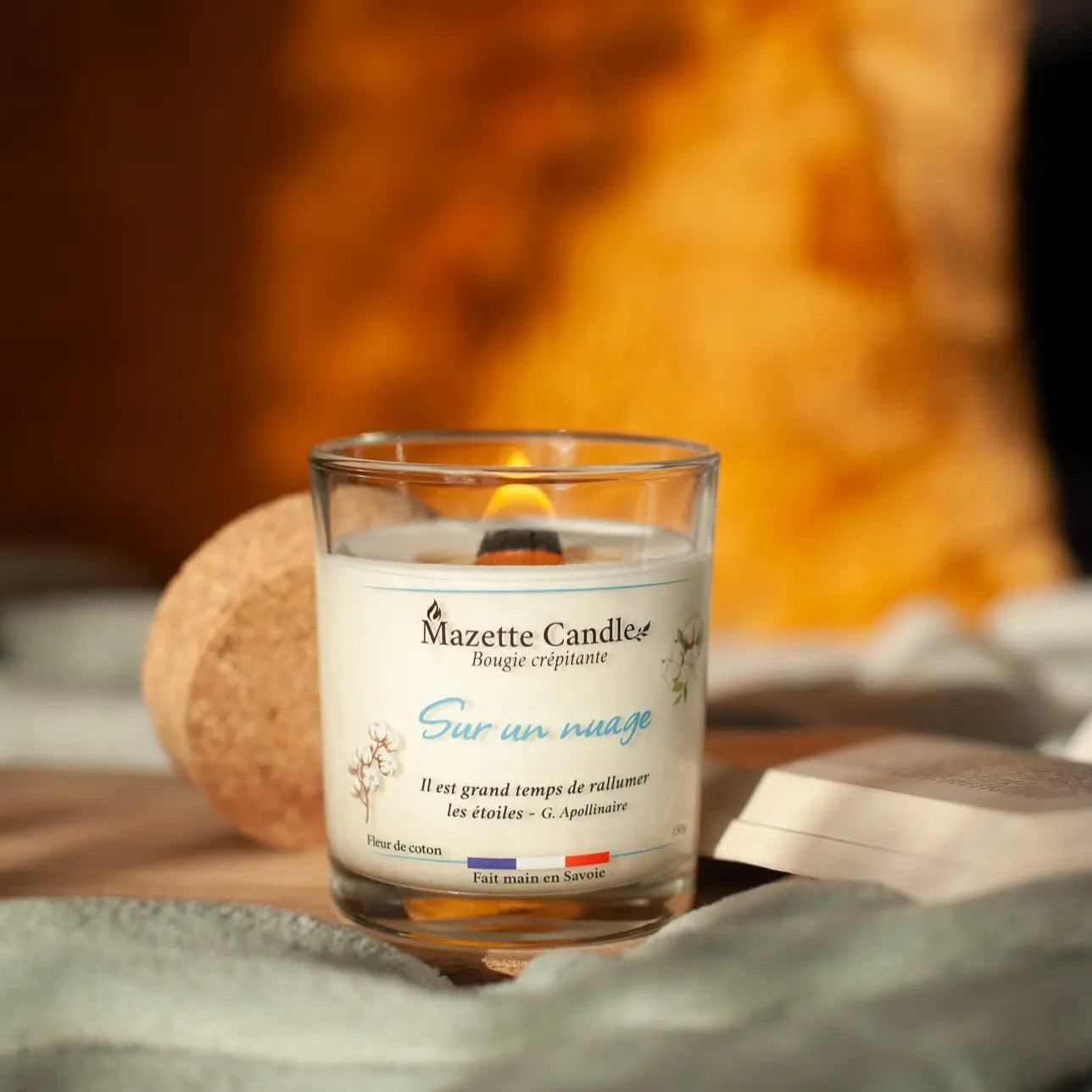 Bougie Mazette Candle Sur un nuage 150g parfum fleur de coton, allumée, avec couvercle en liège et livre dans décor cosy