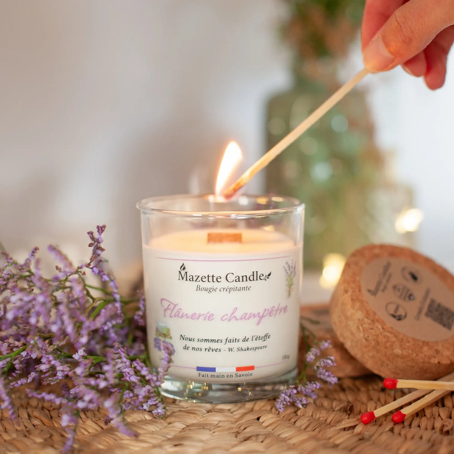 Bougie Mazette Candle Flânerie champêtre 150g parfum lavande, en cours d’allumage avec une allumette, avec couvercle en liège