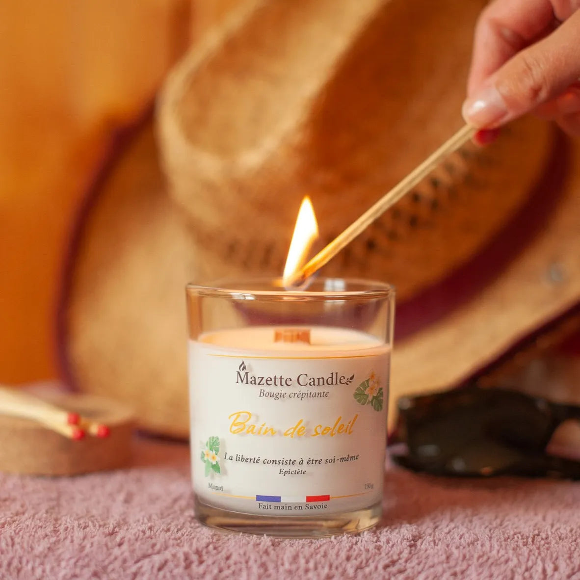 Bougie Mazette Candle Bain de soleil 150g parfum monoï, en cours d’allumage avec une allumette devant un chapeau de paille