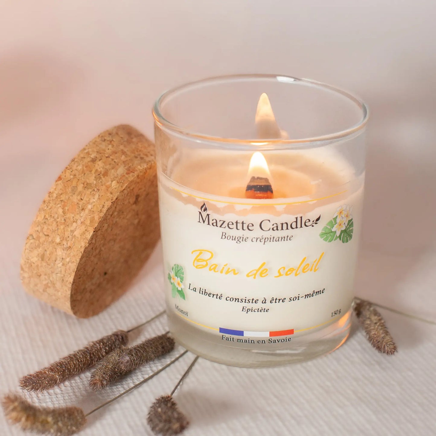 Bougie Mazette Candle Bain de soleil 150g parfum monoï, allumée, avec couvercle en liège à côté et décor d’herbes séchées