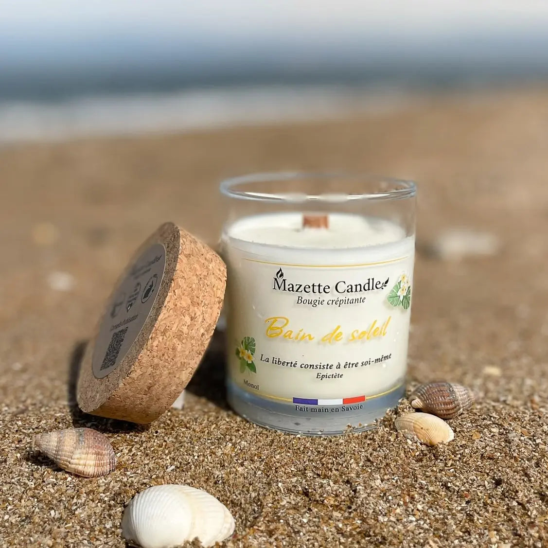 Bougie Mazette Candle Bain de soleil 150g parfum monoï, posée sur le sable avec des coquillages, couvercle en liège à côté