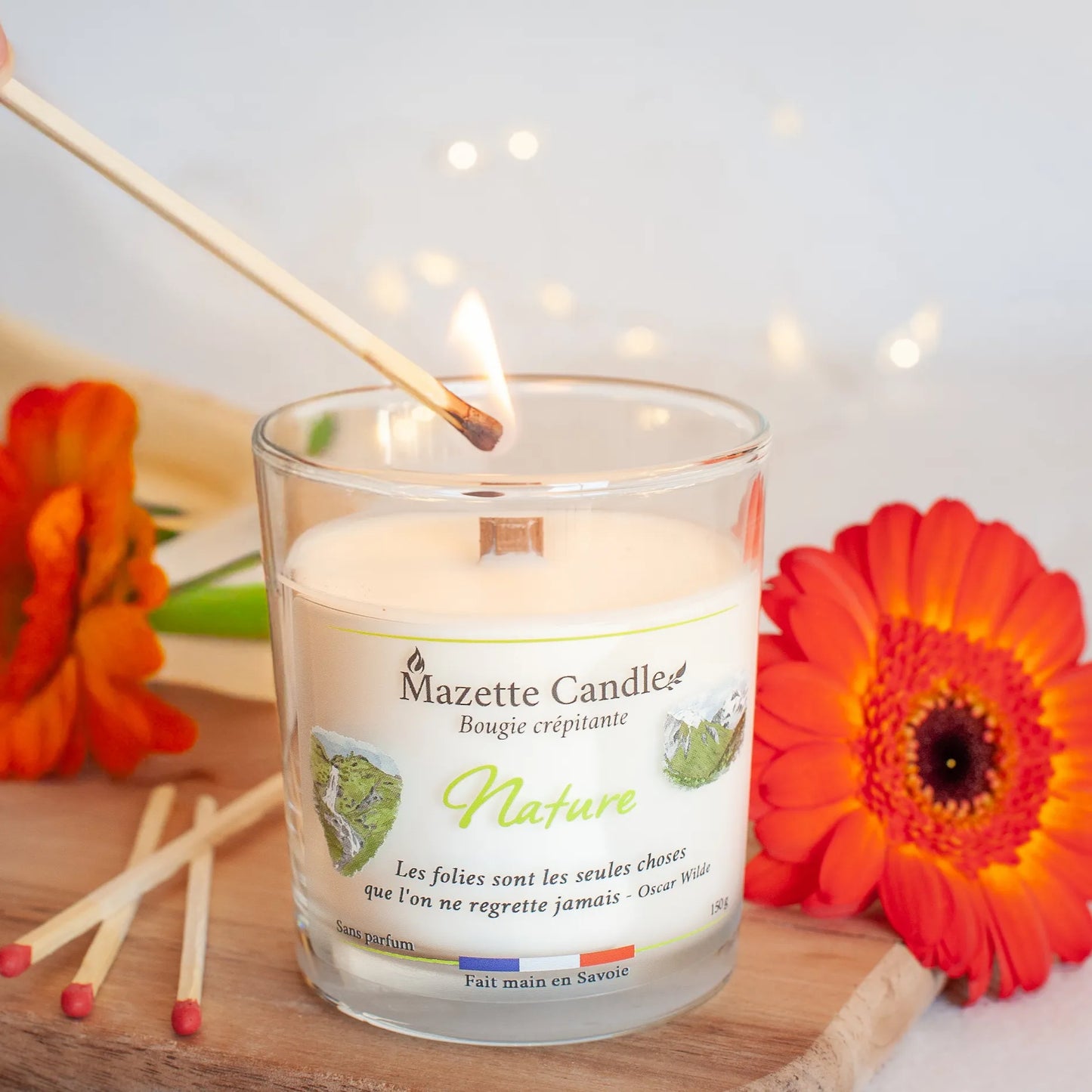 Bougie Mazette Candle Nature 150g sans parfum, en cours d’allumage avec une allumette, avec des fleurs orange en décor
