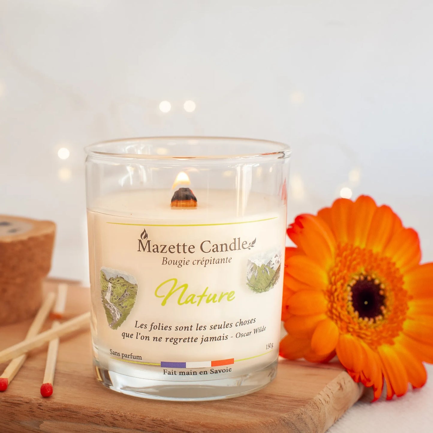 Bougie Mazette Candle Nature 150g sans parfum, allumée et posée sur une planche avec une fleur orange et des allumettes