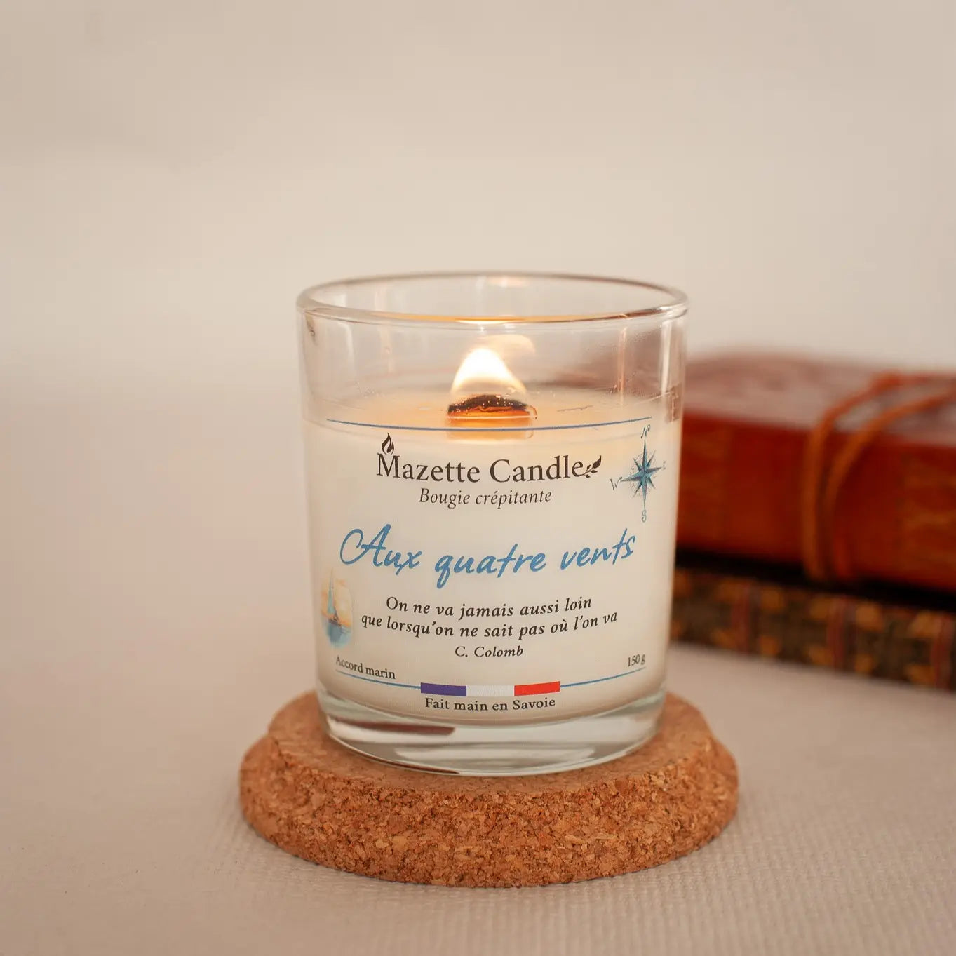 Bougie Mazette Candle Aux quatre vents 150g parfum accord marin, allumée sur un support en liège, carnets de voyage en décor
