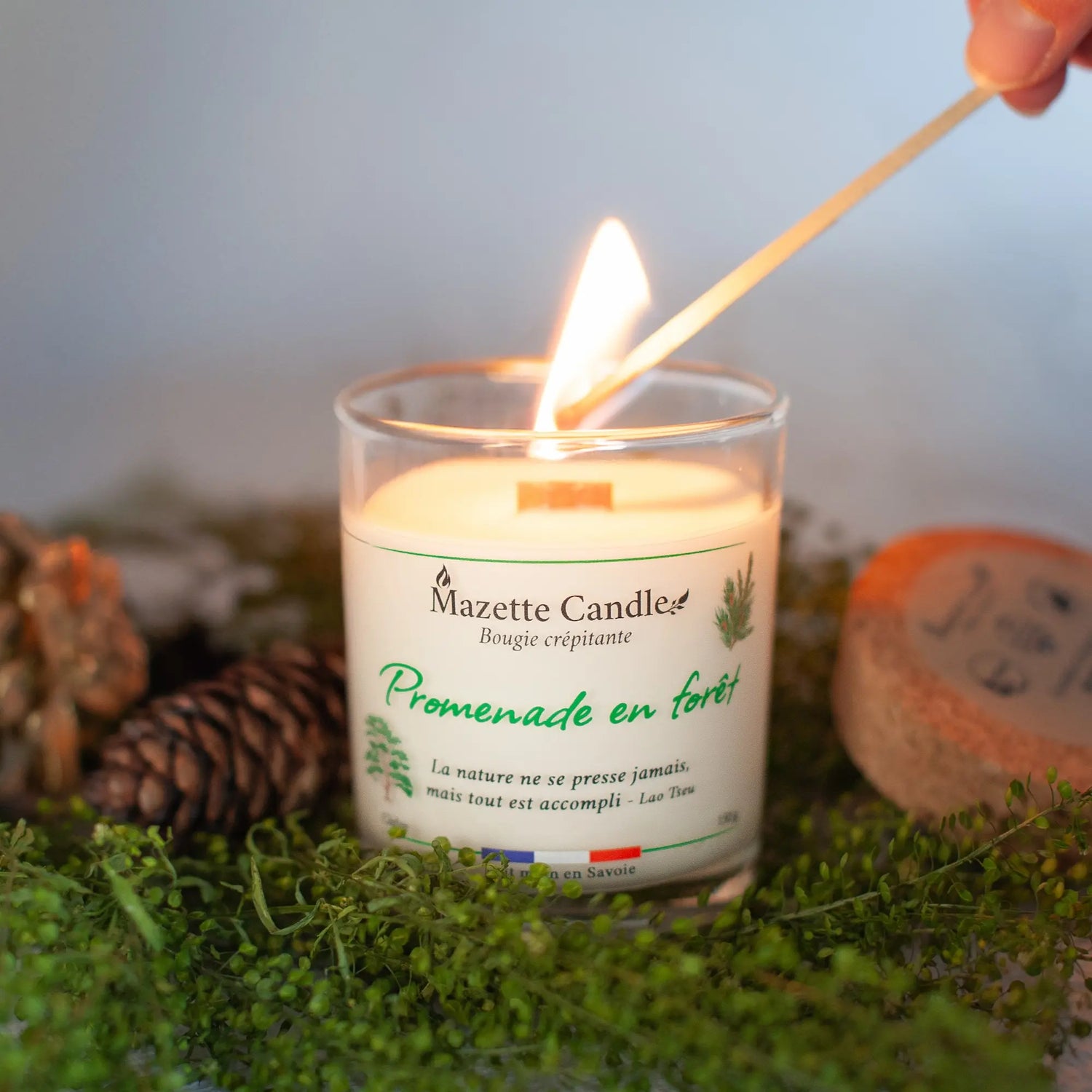 Bougie Mazette Candle Promenade en forêt 150g parfum cèdre, en cours d’allumage avec une allumette, couvercle en liège à côté