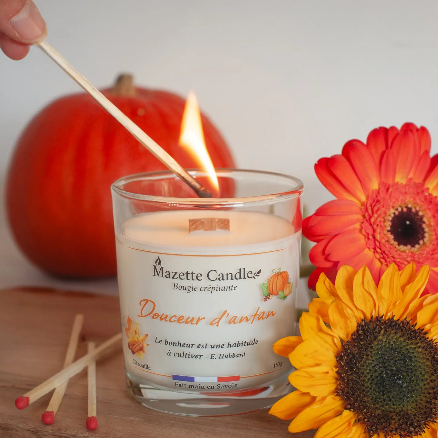 Bougie Mazette Candle Douceur d’antan 150g parfum citrouille, en cours d’allumage avec une allumette, fleurs orange en décor