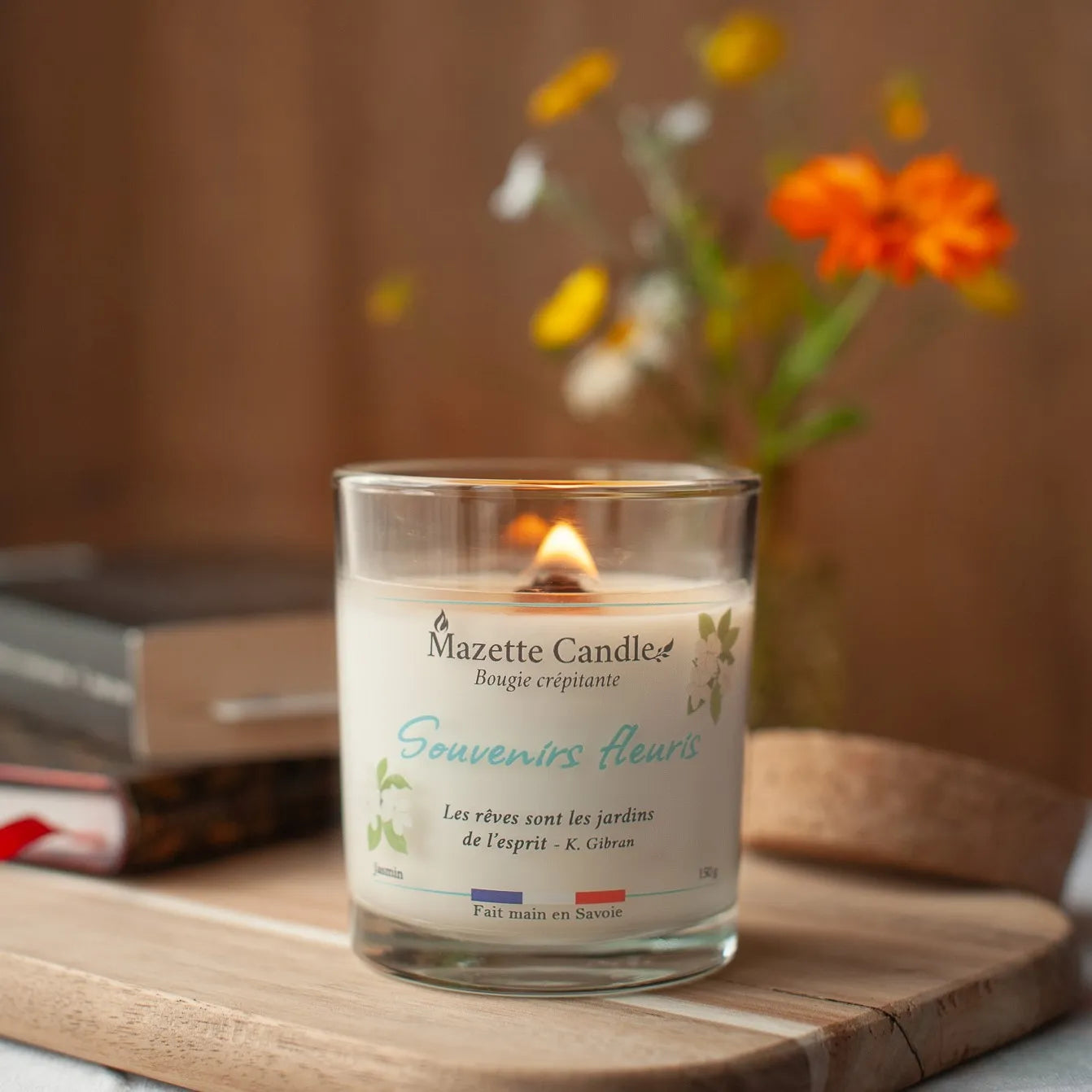 Bougie Mazette Candle Souvenirs fleuris 150g parfum jasmin, allumée devant des livres et un bouquet de fleurs