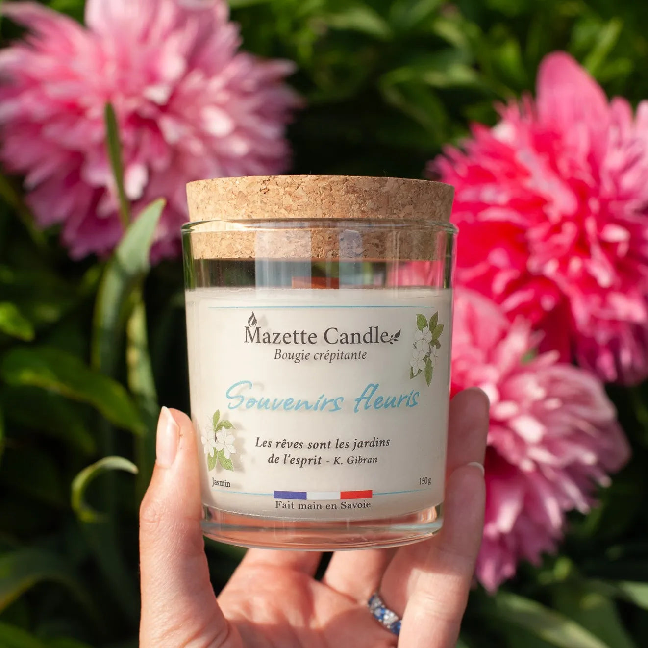 Bougie Mazette Candle Souvenirs fleuris 150g parfum jasmin, tenue dans une main devant des fleurs roses en extérieur