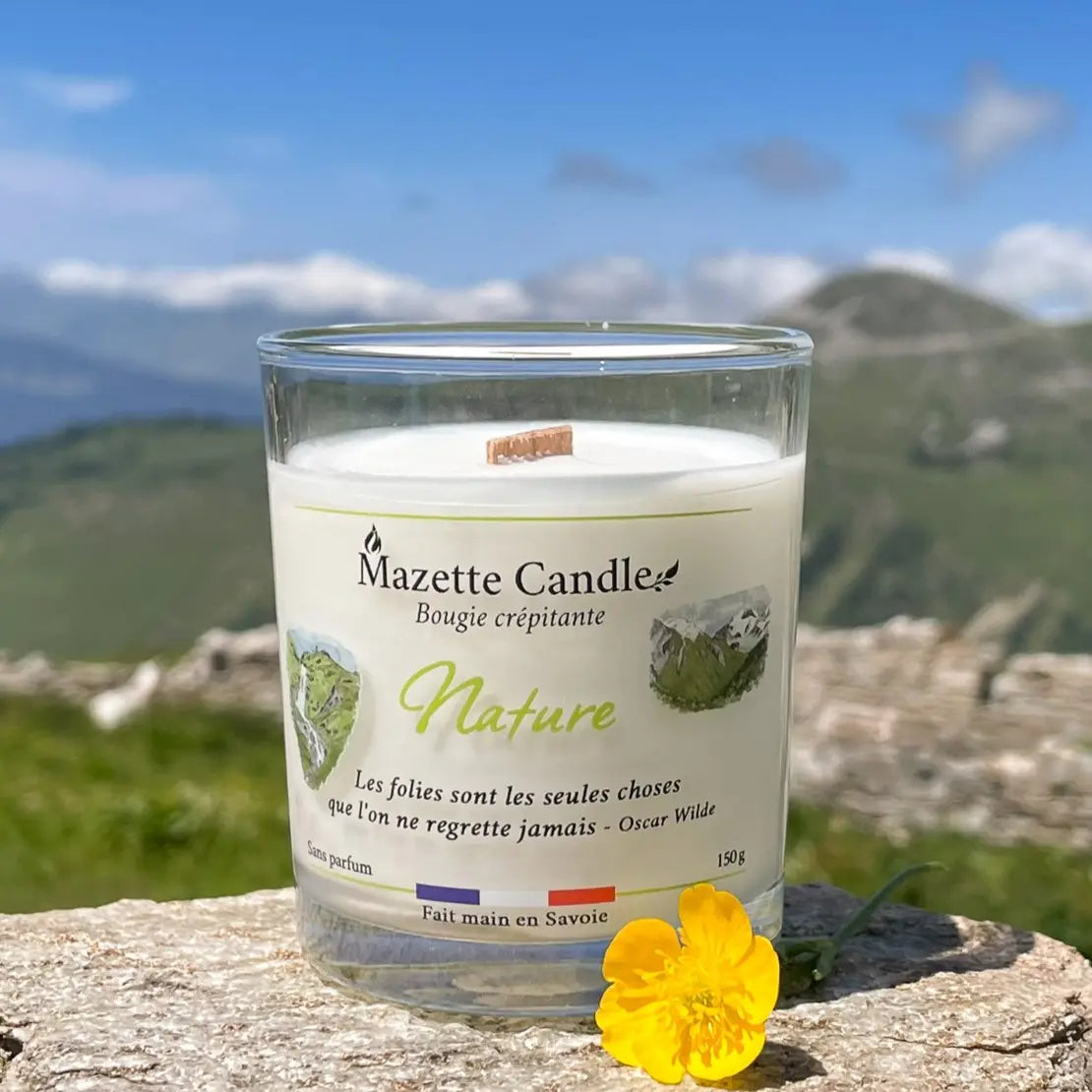 Bougie Mazette Candle Nature 150g sans parfum, posée sur un rocher face aux montagnes, avec une fleur jaune au premier plan