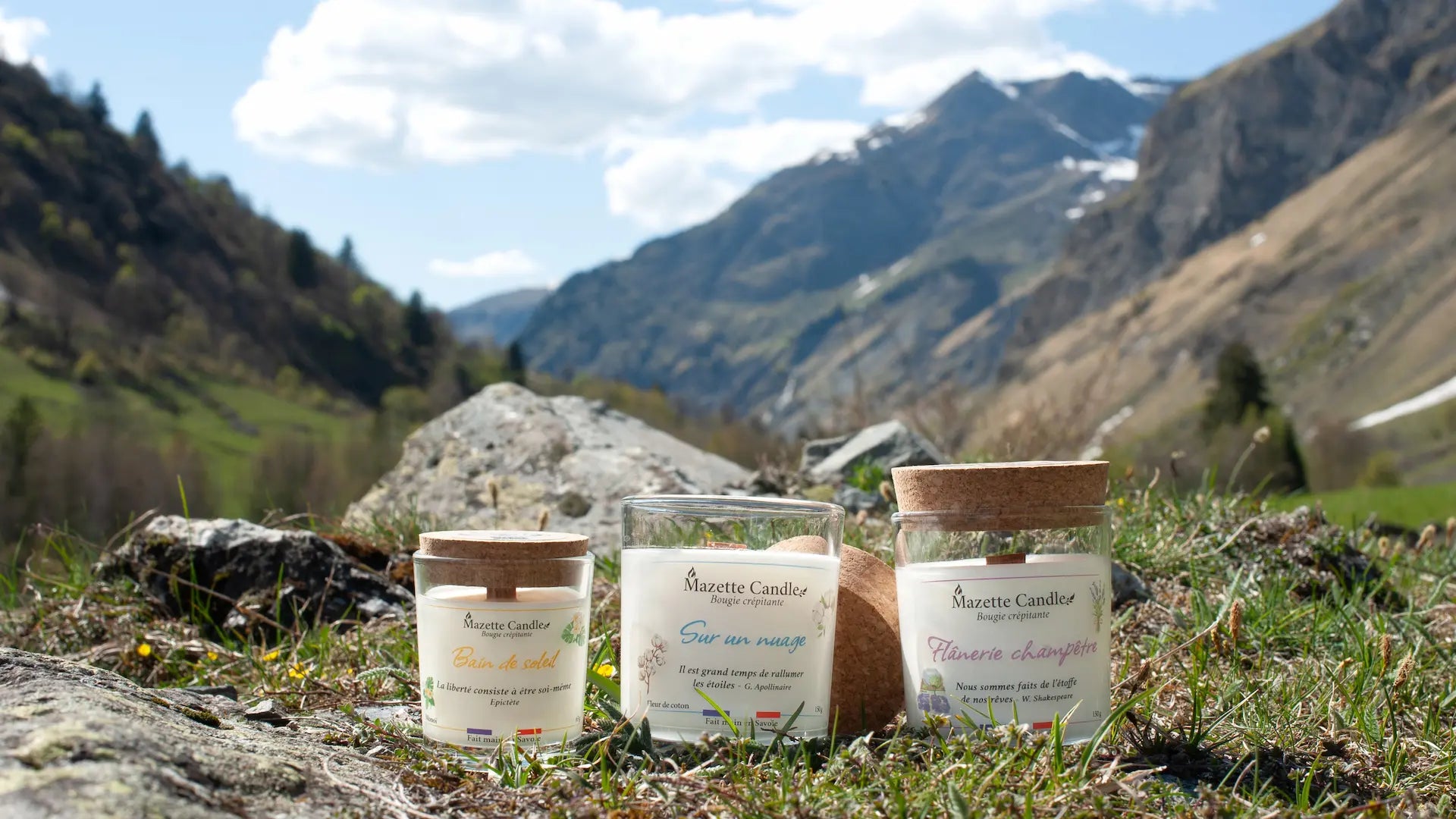 Trois bougies Mazette Candle parfums monoï, fleur de coton et lavande, posées dans l’herbe face aux montagnes alpines
